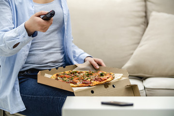 Crop. Unrecognizable woman is sitting on her sofa at home and is watching television while eating pizza. Copy space.