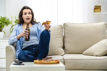 Attractive and interested woman is shocked, watching television. Amazed woman is sitting on the lisht sofa and eating snacks.