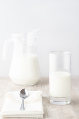 A glass and a jug of milk with a white napkin and metal spoon on a wooden table on white background