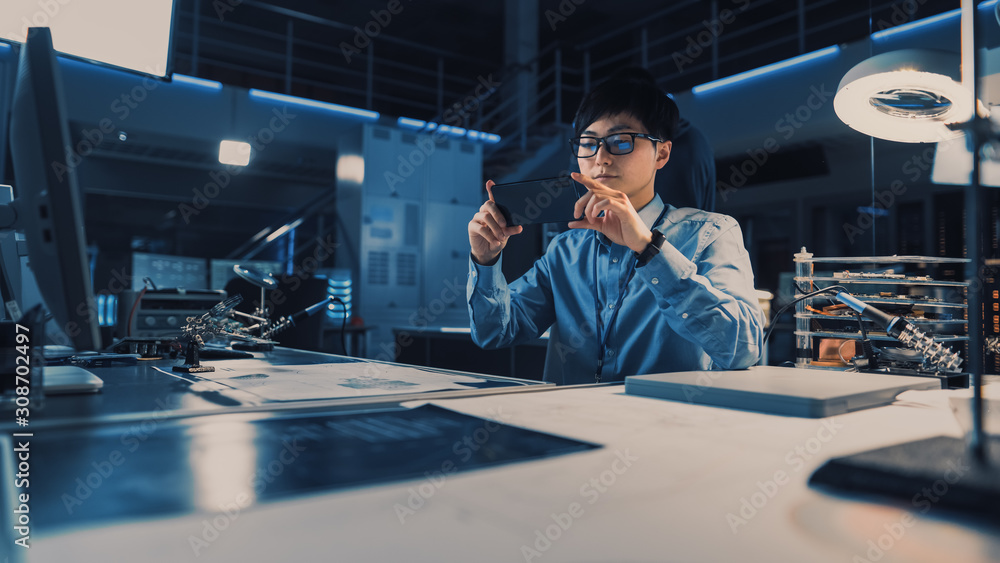 Poster Professional Japanese Development Engineer in Blue Shirt Taking a Photo of Technical Drawings on His Smartpgone in the High Tech Research Laboratory with Modern Computer Equipment.