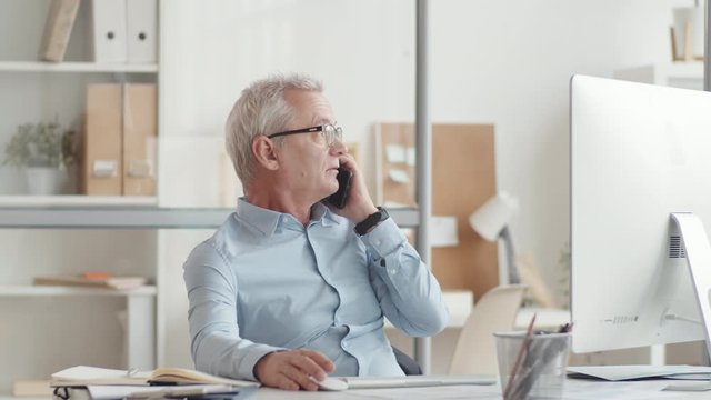 Waist-up Shot Of Senior Caucasian Man With Grey Hair, In Blue Shirt And Glasses, Sitting At Desk In Informal Office, Looking At Computer Screen, Using Mouse And Talking On Smartphone