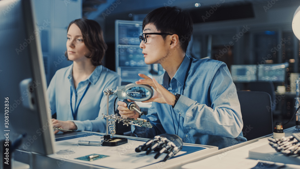 Aufkleber Japanese Development Engineer in Blue Shirt is Soldering a Circuit Board in a High Tech Research Laboratory with Modern Equipment. His Colleague Asks Him a Questio and Points on the Compter Screen.
