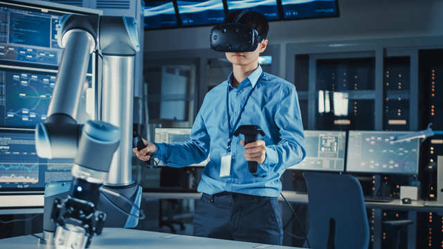 Professional Japanese Development Engineer in Blue Shirt is Controlling a Futuristic Robotic Arm with a Virtual Reality Headset and Joysticks in a High Tech Research Laboratory with Modern Equipment.