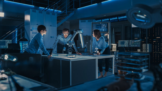 Futuristic Robotic Arm is Moving and Placing a Metal Object. Senior Engineer Talks Through This Advanced Process with His Diverse Team. They are in High Tech Research Laboratory with Modern Equipment