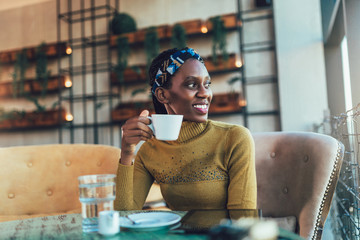 Smiling young African woman drinking a coffee and looking out the window while sitting alone at a table in a cafe