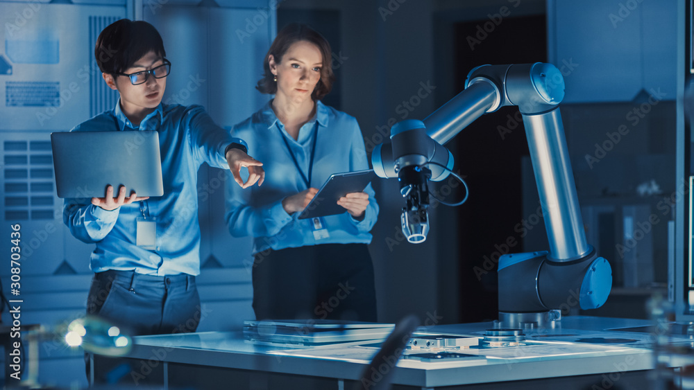 Poster man and woman engineers with laptop and a tablet analyse and discuss how a futuristic robotic arm wo