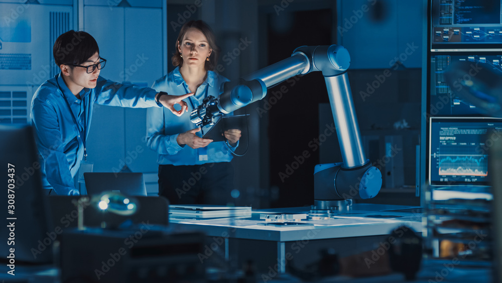 Fototapete man and woman engineers with laptop and a tablet analyse and discuss how a futuristic robotic arm wo