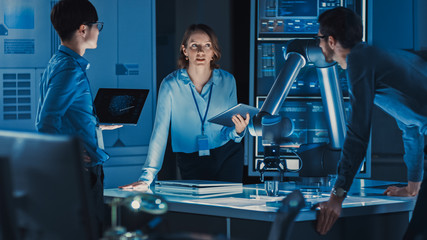 Diverse Team of Engineers with Laptop and a Tablet Analyse and Discuss How a Futuristic Robotic Arm...