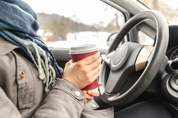Driver drinking coffee in the car, driving and holding a steering wheel and cup of hot beverage at cold winter day with snow behind window