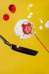 Fried egg and frying pan with a red watch, paint jars and spray paints and a brush paint levitate in the air.