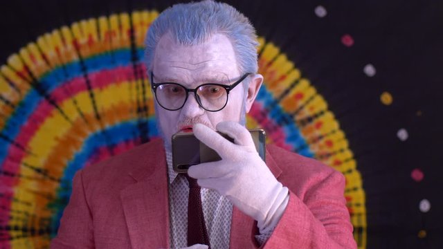 elderly mime man in big glasses with blue hair and beard in bright pink jacket and tie looks at smartphone screen and shows it to audience. Laughter, tears, positive and negative emotions. Happy clown