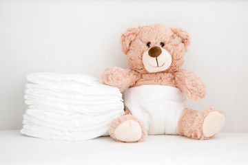 Brown teddy bear sitting in baby bed. Stack of white diapers. Front view. Closeup. - obrazy, fototapety, plakaty