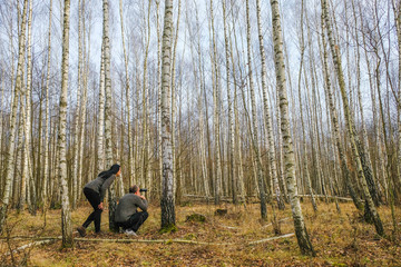 Happy couple in identical gray sweaters watching binoculars in birch forest in Ukraine. The man sat down. The woman leaned toward him. Rest together.
