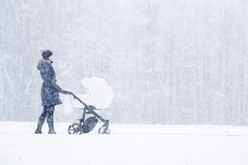 Young alone mother pushing white baby stroller. Woman walking in heavy blizzard at park. Winter day...