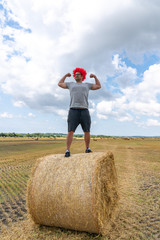 Strong young man in red wig stands on the round haystack among the field and shows his biceps