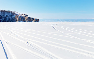 Lake Baikal on a sunny winter day. Car tracks on the snowy ice of a lake. Near the cliff, a group of tourists on a picnic. Winter ice travel concept