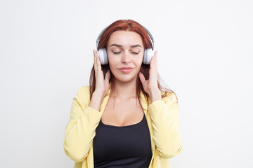 Fototapeta premium A red haired girl in a yellow business suit listens to music in large headphones on a white background
