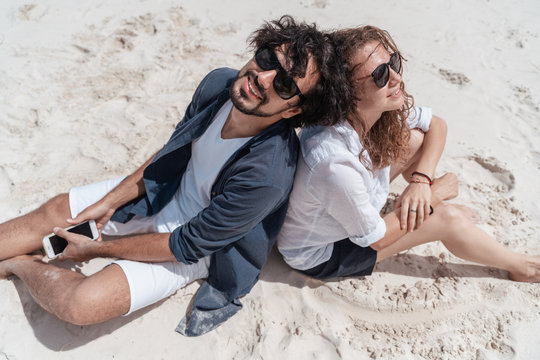 Beautiful young couple joyfully sitting on the white sand by the sea and takes a selfie on a smartphone. Honeymoon Travel and Vacation on the Tropical Ocean.