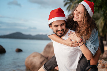 Obraz premium Young beautiful couple in Santa Claus hats walks along the shore of the tropical sea at the resort, vacation and travel for the New Year and Christmas time.
