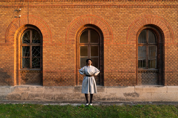 Young girl in grey dress and black beret stands near the red brick wall, holds her hands on her waist