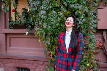 Happy smiling young brunette female in glasses, dressed in plaid jacket and white shirt, stands near the red wall with green ivy