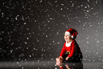Laughing infant child baby boy toddler is sitting in santa claus costume and cap under the snow on dark with copy space