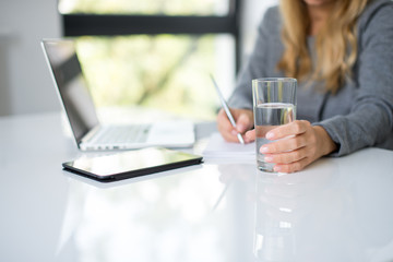 A blonde hair businesswoman working in her workstation. Businesswoman working at the desk at workplace. Woman working in home office. Woman sitting at desk and working with laptop and glass of water.