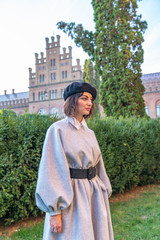 Side view portrait of the attractive brunette girl in grey dress and black beret stands near the green bushes, old vintage building on the background