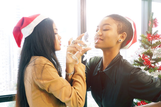 Two Attractive And Beautiful Asian And African Women With Their Arms Linked Drinking Wine In Christmas And New Year Party Celebration In The Office. Colleagues Having Fun And Good Time Concept