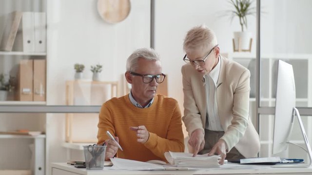 Medium shot of grey-haired Caucasian male business owner sitting at desk in office, female secretary bringing thick pile of documents and leaving, director looking at it then grabbing head in despair
