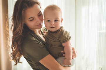 Home portrait of a baby boy with mother near the window. Mom holding and kissing her child.