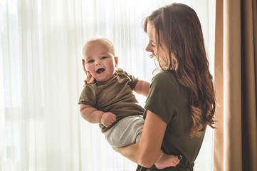 Home portrait of a baby boy with mother near the window. Mom holding and kissing her child.