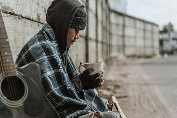Homeless man sitting on the street in the shadow of the building and begging for help and money. Problems of big modern cities.