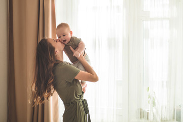 Home portrait of a baby boy with mother near the window. Mom holding and kissing her child.