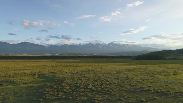 Drone takes off above wide green valley toward mountain snowy peaks at sunset