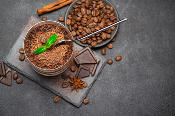 Classic tiramisu dessert in a glass cup and pieces of chocolate on stone cutting board on darl concrete background