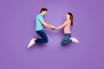 Fototapeta na wymiar Full size profile photo of amazed two people guy lady jumping high holding hands rejoicing having fun wear casual blue striped t-shirts jeans footwear isolated purple color background