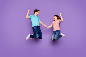 Full body photo of funny two people guy lady jumping high holding hands rejoicing ecstatic achievement wear casual blue striped t-shirts jeans footwear isolated purple color background