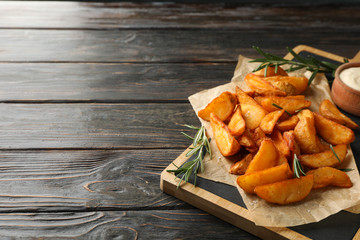 Slices of baked potato wedges, rosemary, white sauce, kitchen board on wooden background, space for text. Closeup