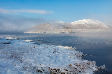 Snowy shore of mountain lake with thin ice and fog over it