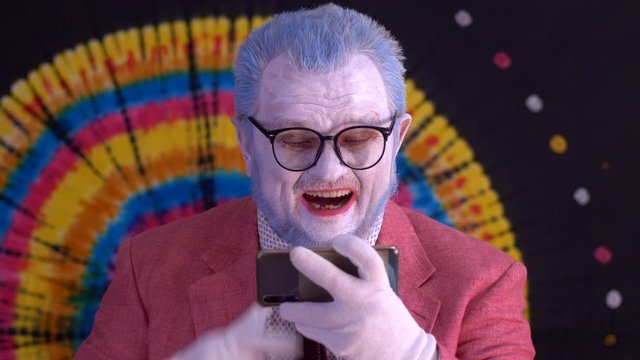 elderly mime man in big glasses with blue hair and beard in bright pink jacket and tie looks at smartphone screen and shows it to audience. Laughter, tears, positive and negative emotions. Happy clown