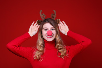Studio shot of beautiful smiling young woman wearing reindeer horns headband for Christmas. Attractive blonde female in red turtleneck sweater, new years eve. Copy space, isolated background, close up