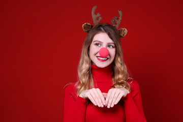 Studio shot of beautiful smiling young woman wearing reindeer horns headband for Christmas. Attractive blonde female in red turtleneck sweater, new years eve. Copy space, isolated background, close up