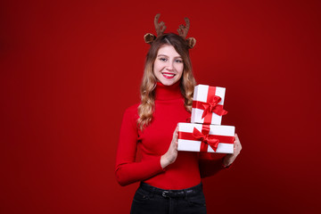 Fototapeta premium Studio shot of beautiful smiling young woman wearing reindeer horns headband for Christmas. Attractive blonde female in red turtleneck sweater, new years eve. Copy space, isolated background, close up