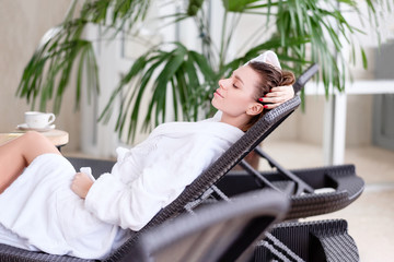 Young beautiful relaxed positive woman with eyes closed in white coat relaxing on a couch at a hotel. Concept of wellness and rehabilitation procedures