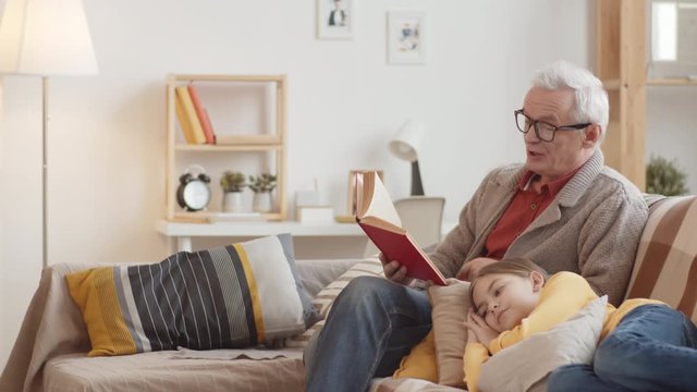 Tilting full shot of elderly Caucasian grandfather with grey hair in glasses and cardigan sitting on couch and reading aloud book, and young granddaughter lying on pillows and listening