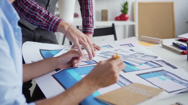 Tilt down closeup of man explaining ideas to colleague at desk in office. Two web designers or marketing professionals discussing website interface samples for different devices printed on paper
