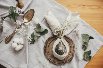 Stylish Easter brunch table setting with egg in easter bunny napkin. Modern natural dyed gray egg on napkin with bunny ears, flowers on vintage plate and cutlery. Easter table decorations