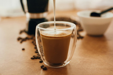 Pouring milk into coffee in glass cup on background of roasted coffee beans, grounded coffee,...