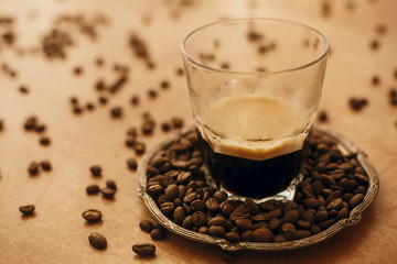 Hot espresso with foam in glass cup on vintage plate with aromatic roasted coffee beans on wooden table. Copy space. Alternative Coffee brewing concept.